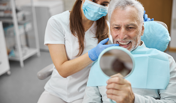 Older man sitting in a dental chair looking in a handheld mirror at his new dental implants from Rocket City Prosthodontics in Huntsville, AL