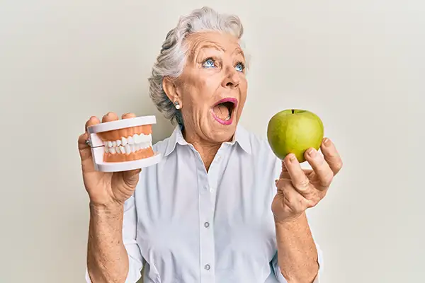 Elderly woman excitedly holding a set of dentures in one hand and a green apple in the other, symbolizing the comfort and functionality of dentures.