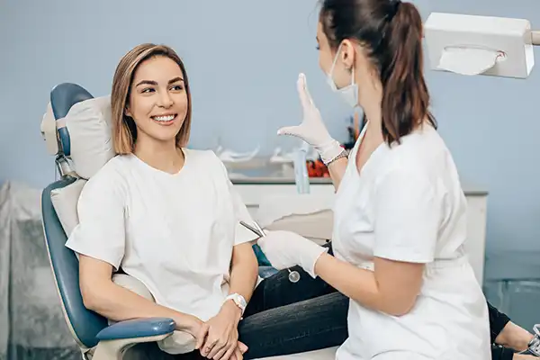 A smiling patient sits in a dental chair and listens while the dental hygienist welcomes her and discusses her first visit.